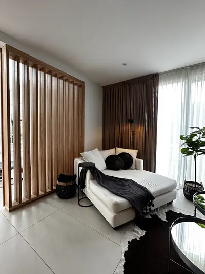 Chaise longue blanca junto a un divisor de listones de madera y un ventanal con cortinas.